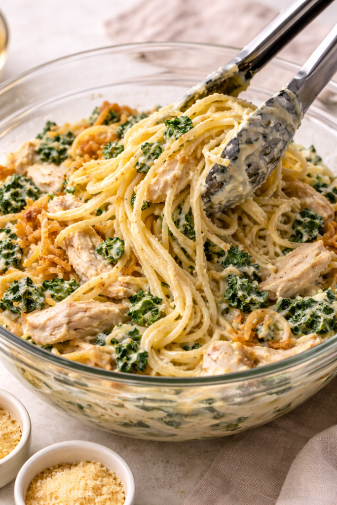 Spaghetti being tossed with rotisserie chicken, spinach, cheese, and creamy sauce in a large glass bowl with tongs