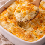 Wooden spoon scooping creamy party potatoes from a baked mashed potato casserole with melted cheddar, buttery breadcrumb topping, and chives on a marble countertop.