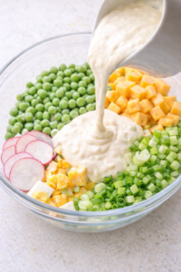 Creamy dressing being poured over green peas, cheddar cheese, eggs, celery, and radishes in a clear glass bowl.