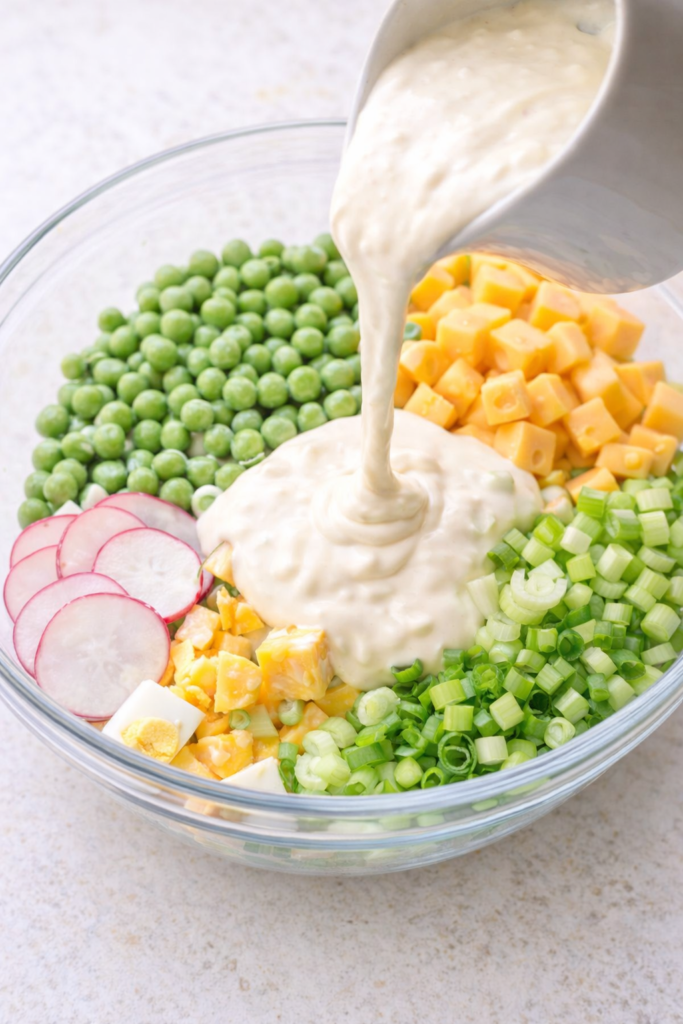 Creamy dressing being poured over green peas, cheddar cheese, eggs, celery, and radishes in a clear glass bowl.