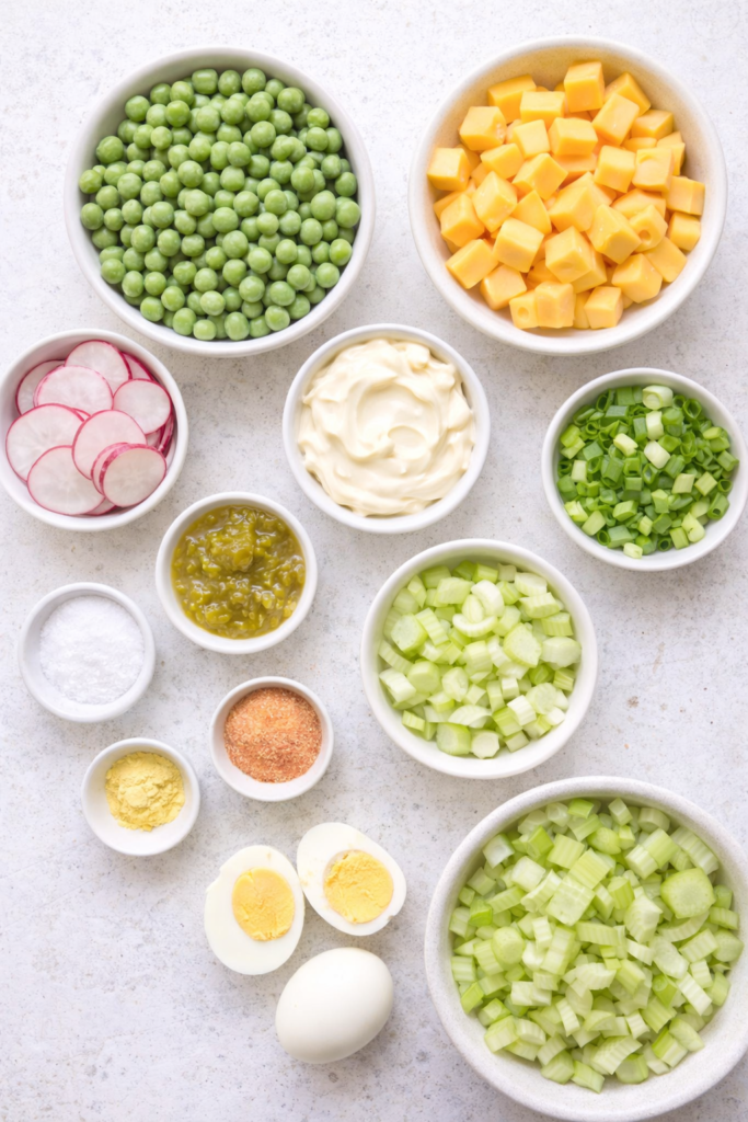 Flat lay of ingredients for classic pea and cheese salad including green peas, cheddar cheese, celery, radishes, eggs, and creamy dressing.