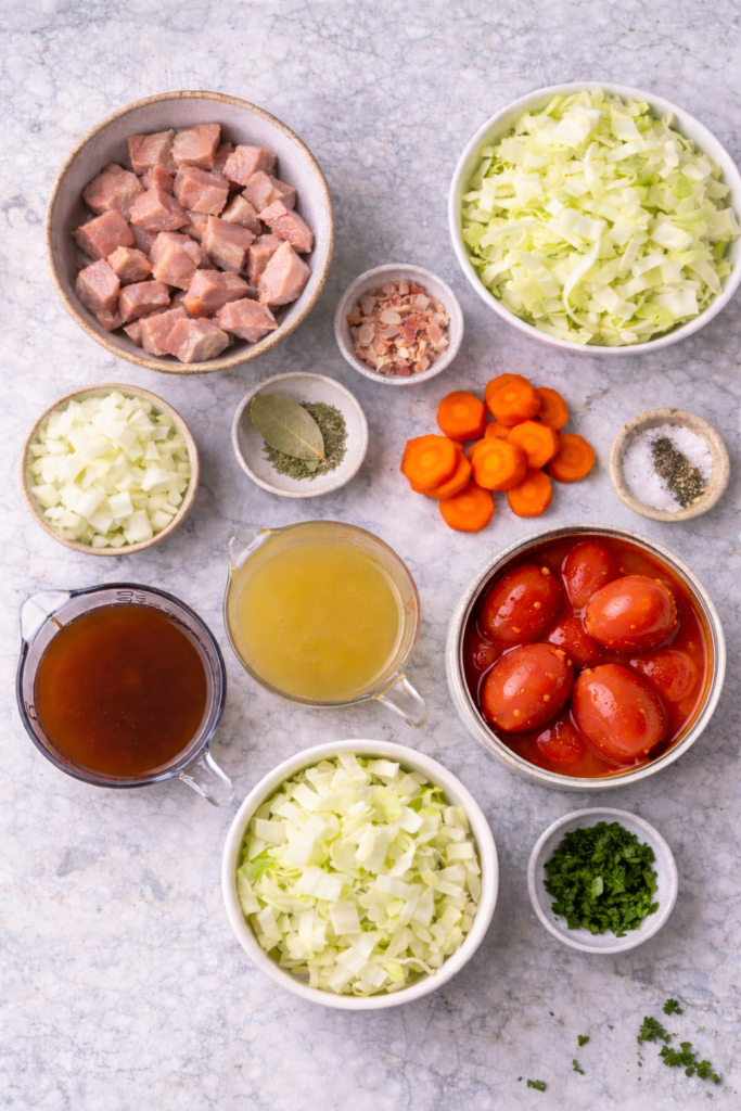 Flat lay of pork and cabbage soup ingredients including raw pork, chopped cabbage, carrots, onion, bacon, broths, herbs, and a can of whole tomatoes on a marble surface