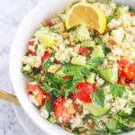 Riced cauliflower tabbouleh salad with fresh herbs, cherry tomatoes, cucumber, and lemon served in a white bowl on a marble table.