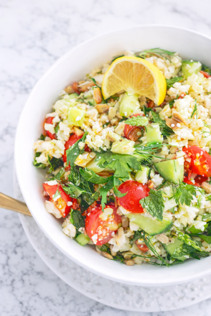 Riced cauliflower tabbouleh salad with fresh herbs, cherry tomatoes, cucumber, and lemon served in a white bowl on a marble table.