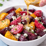 Roasted multicolor beets cut into wedges in a white bowl with cider vinaigrette being poured over and crumbled blue cheese on top