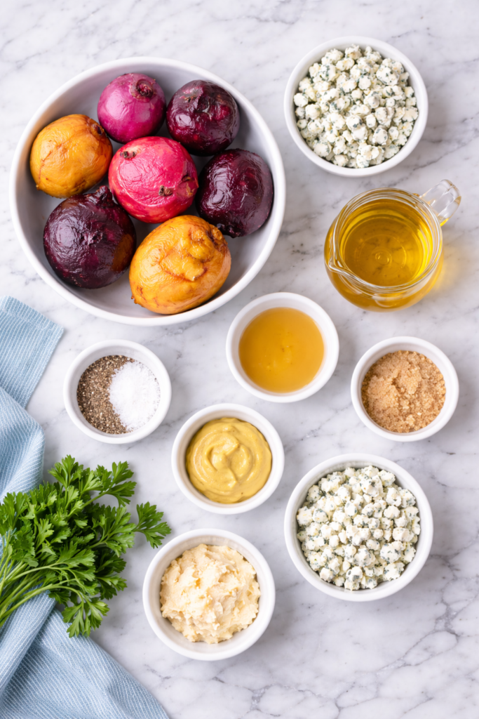 Flat lay of ingredients for roasted beet salad including multicolor beets, olive oil, apple cider vinegar, Dijon mustard, horseradish, brown sugar, blue cheese, and fresh parsley on a marble surface