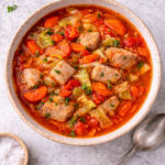 Bird’s-eye view of rustic pork and cabbage soup with carrots and tomatoes in a ceramic bowl on a light marble background