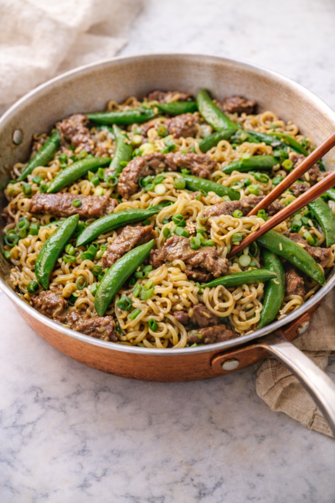 Sirloin snap pea stir fry with ramen noodles cooking in a copper skillet on a marble countertop, garnished with green onions.