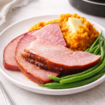Sliced brown sugar glazed ham served with mashed sweet potatoes and green beans on a white plate with whole ham roast in background.