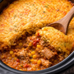 Vertical image of homemade tamale pie cooked in a crockpot with a serving scooped out, showing layers of tender pork, corn, and tomato filling beneath a golden cornbread crust.