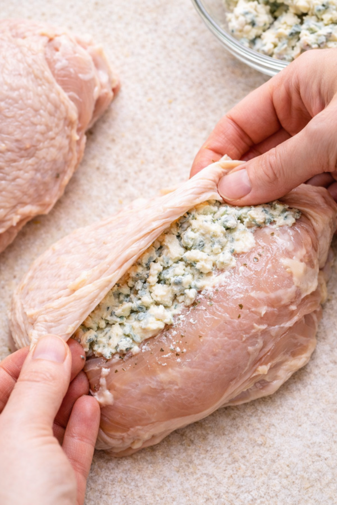 Hands stuffing blue cheese butter under the skin of a raw chicken breast before baking