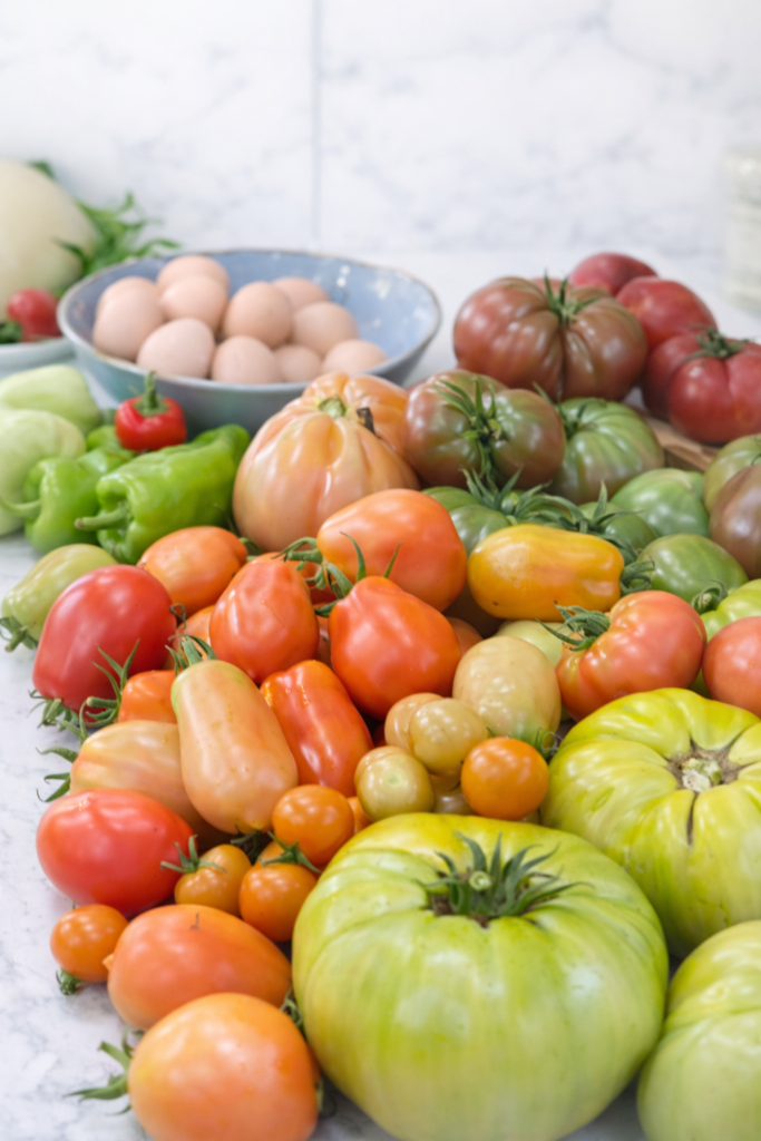 Assorted heirloom and cherry tomatoes from a summer garden harvest arranged on a marble countertop with fresh eggs and peppers, ready for canning and preserving.