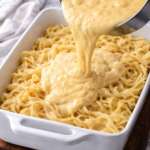 Creamy Swiss cheese sauce being poured over homemade egg noodles in a white casserole dish for a German noodle bake.