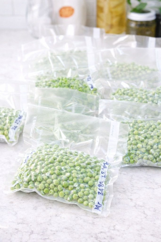 Vacuum-sealed bags of fresh green peas stacked on a white countertop with a food saver and basket of pea pods in the background.