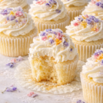 Vanilla cupcakes topped with swirled buttercream and colorful candied edible flower petals, styled close together on a light neutral surface and sized vertically for Pinterest.