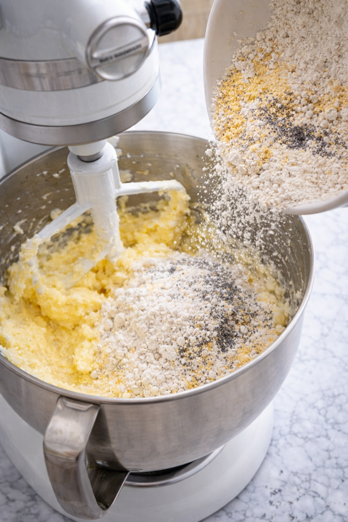 Dry ingredients including flour, cornmeal, and poppy seeds being added to creamed butter and sugar in a stand mixer for lemon poppy seed cornmeal cake