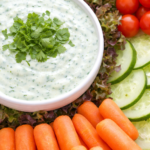 Creamy Asiago cilantro garlic yogurt dip in a white bowl served with fresh vegetables including carrots, cucumbers, cherry tomatoes, and lettuce.