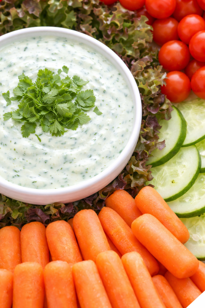 Creamy Asiago cilantro garlic yogurt dip in a white bowl served with fresh vegetables including carrots, cucumbers, cherry tomatoes, and lettuce.