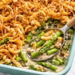 Creamy asparagus casserole topped with crispy fried onions being scooped with a spoon from a baking dish on a marble countertop