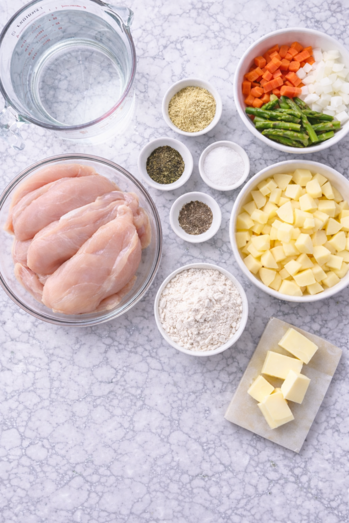 Flat lay of ingredients for asparagus chicken chowder including chicken breasts, potatoes, carrots, onion, asparagus, butter, flour, and seasonings on marble counter