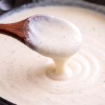 thick béchamel sauce coating a wooden spoon showing smooth creamy white sauce texture in skillet