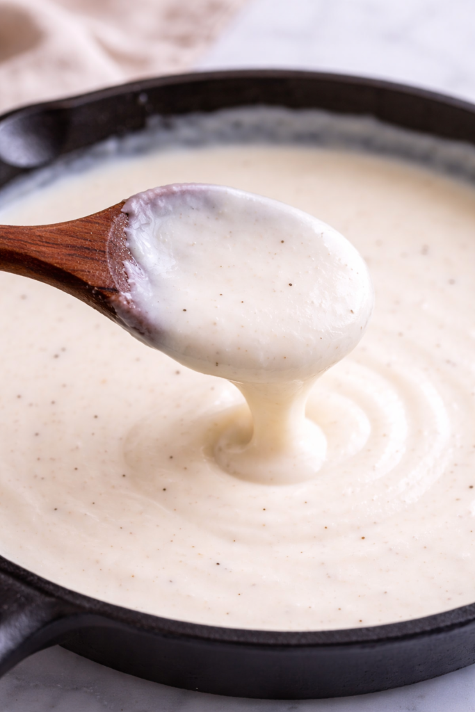 thick béchamel sauce coating a wooden spoon showing smooth creamy white sauce texture in skillet