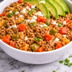 Beefy Spanish rice skillet in a white bowl topped with sliced avocado and crumbled queso fresco on a marble counter