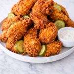 Crispy fried chicken piled on a white platter with sliced pickles and ranch