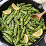 Blistered sugar snap peas in a black cast iron skillet with lemon slices and garlic aioli dipping sauce