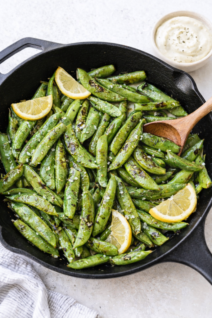 Blistered sugar snap peas in a black cast iron skillet with lemon slices and garlic aioli dipping sauce