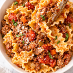 tongs serving mafalde pasta with homemade bolognese sauce into white bowl on marble counter