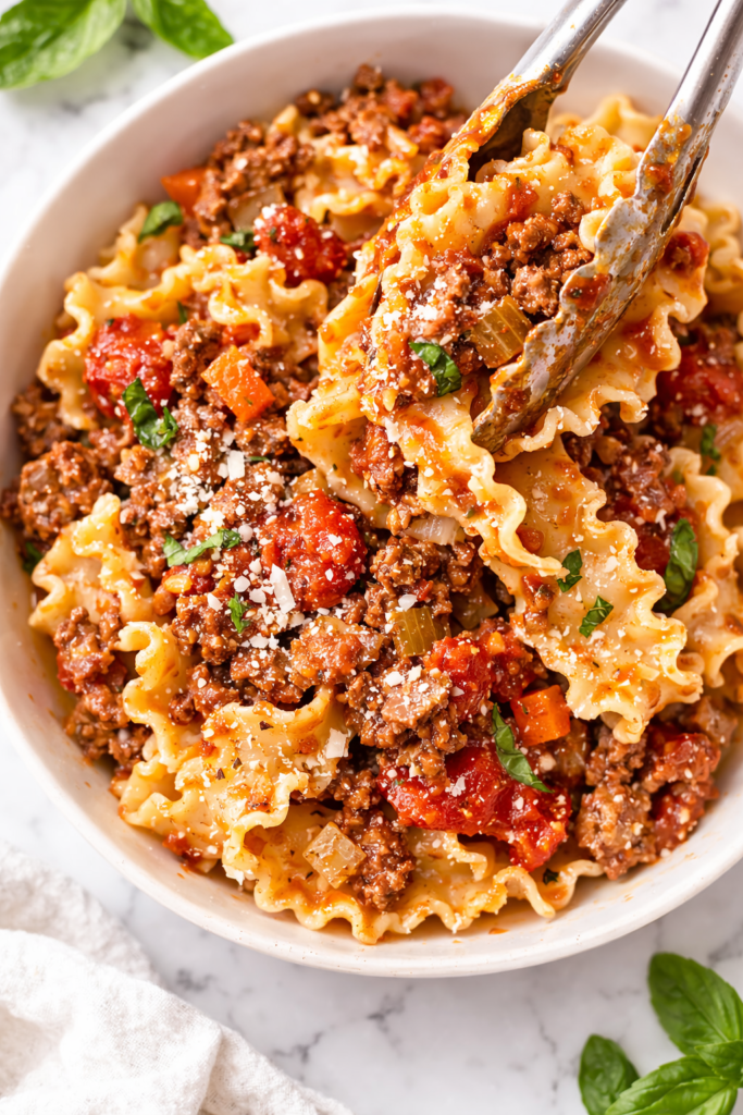 tongs serving mafalde pasta with homemade bolognese sauce into white bowl on marble counter