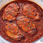 Boneless pork chops simmering in smooth rhubarb barbecue sauce in a white Dutch oven on a marble counter