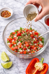 Making Brazilian tomato onion vinaigrette in a glass bowl with diced tomatoes, onions, parsley and vinegar on a marble countertop.