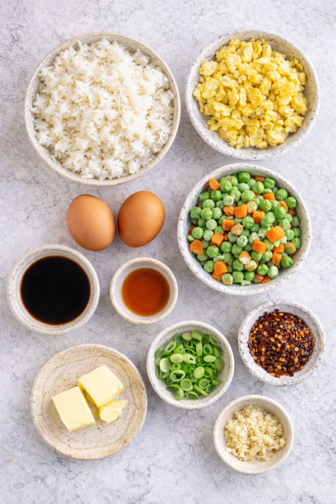 Flat lay of ingredients for budget egg fried rice including cooked rice, eggs, peas and carrots, soy sauce, sesame oil, green onions, butter, garlic, and chili crisp on a marble surface.