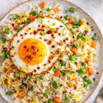 Budget egg fried rice topped with a crispy fried egg and chili crisp on a handmade pottery plate over a marble countertop.