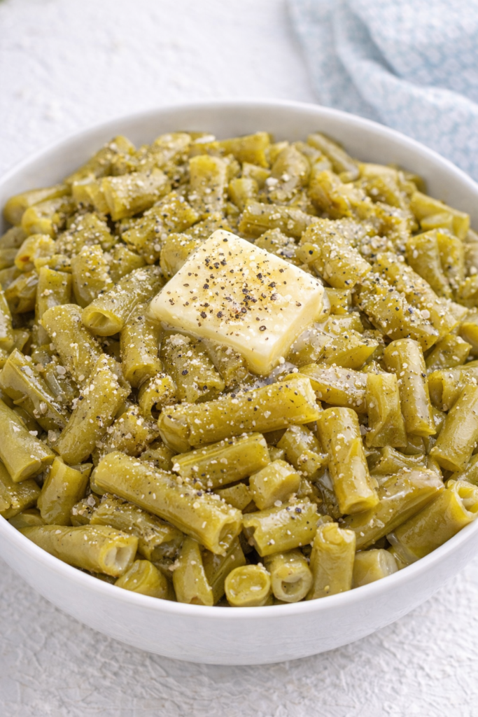 Canned green beans in a white bowl with butter melted on top and sprinkled salt and pepper
