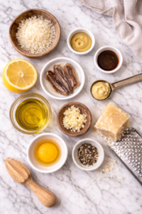 Flat lay of Caesar dressing ingredients on a marble countertop including egg yolk, anchovies, garlic, lemon, olive oil, Dijon mustard, Worcestershire sauce, Parmesan, and black pepper.