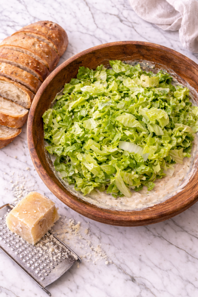 Romaine lettuce sitting on top of Caesar dressing in a wooden bowl before tossing with Parmesan cheese and crusty bread on a marble countertop.