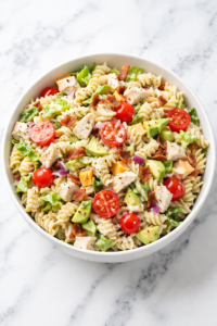 California chicken club pasta salad in a white bowl with bacon, chicken, avocado, and tomatoes on a marble countertop