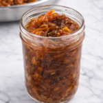 Jar of golden brown caramelized onions on a marble countertop with a skillet of onions in the background
