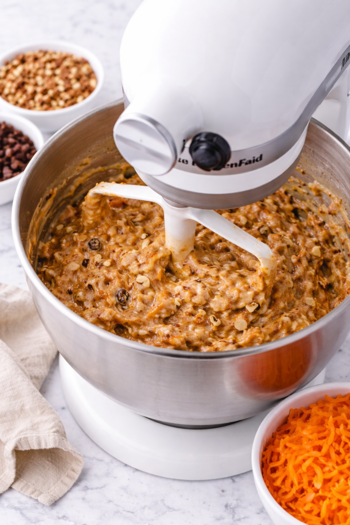 White stand mixer mixing carrot cake cupcake batter in a stainless bowl on a marble counter with shredded carrots, pecans, and raisins nearby.