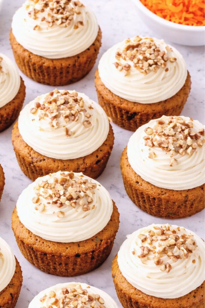 Close-up overhead view of frosted carrot cake cupcakes topped with chopped pecans on a white marble counter.