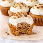 Carrot cake cupcakes with cream cheese frosting and chopped pecans on top, with one cupcake bitten open on wrinkled parchment paper over a white marble counter.