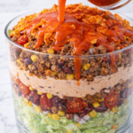 Catalina dressing being poured over layered taco salad with lettuce, beans, corn, seasoned beef, tomatoes, and Doritos in a glass bowl on a marble countertop