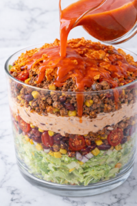 Catalina dressing being poured over layered taco salad with lettuce, beans, corn, seasoned beef, tomatoes, and Doritos in a glass bowl on a marble countertop