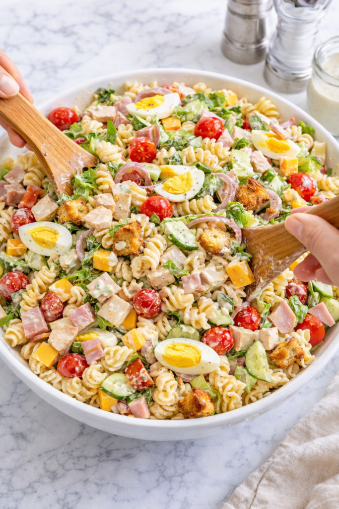 chef salad pasta salad being tossed with wooden spoons rotini pasta ham turkey cheese eggs and creamy dressing in white bowl