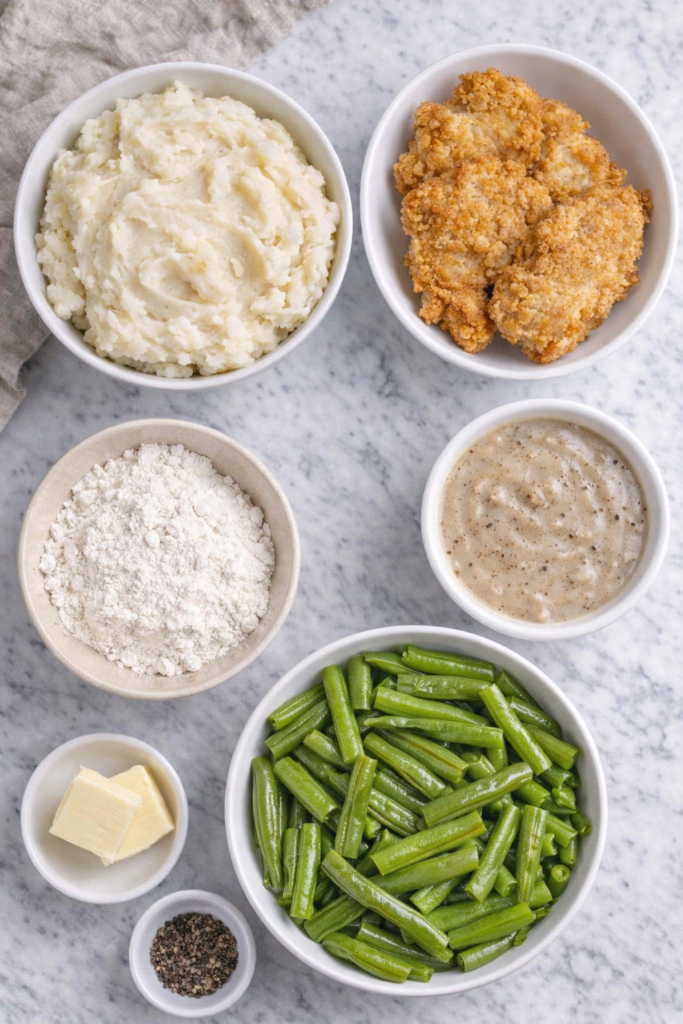 Flat lay of ingredients for chicken fried steak mashed potato bowls including mashed potatoes, chicken fried steak, green beans, pepper gravy, flour, butter, and black pepper on a marble countertop