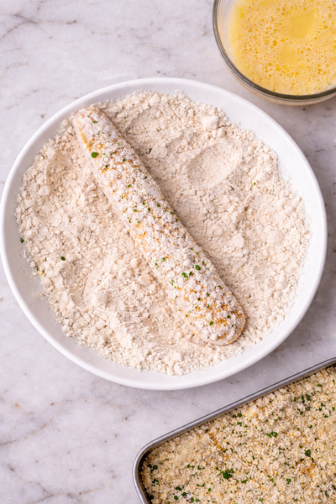 raw chicken strip coated in flour on a white plate during chicken fries breading process