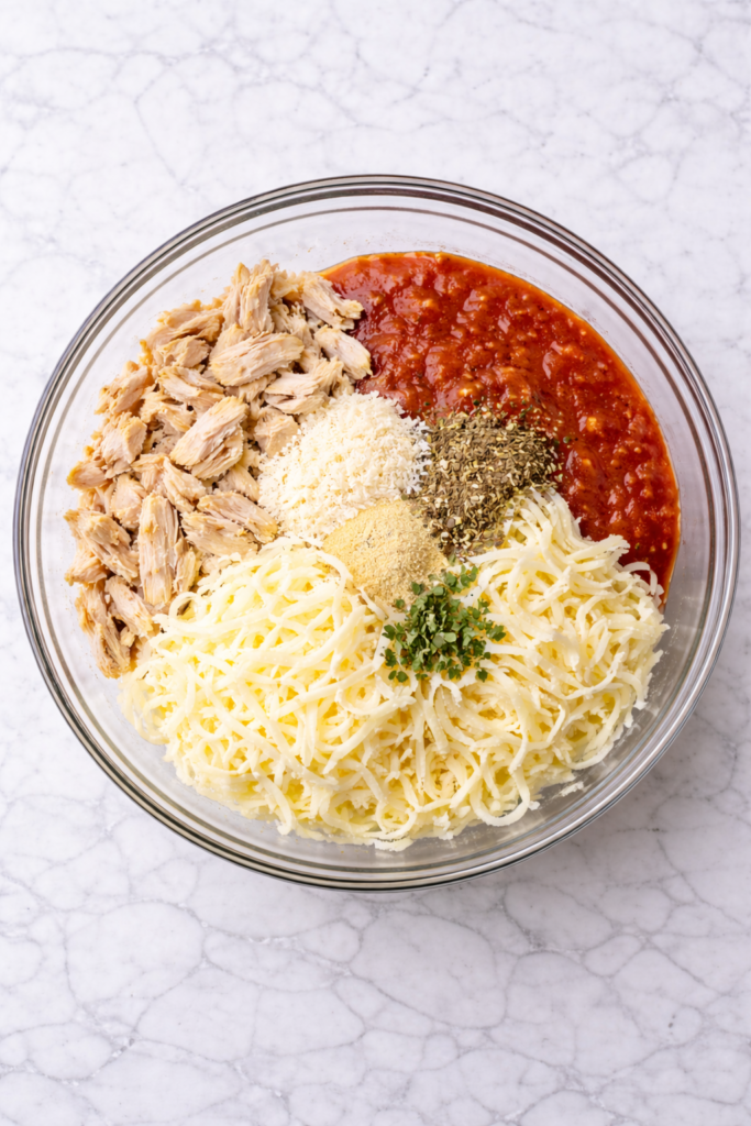 Chicken parmesan egg roll filling ingredients in a glass bowl before mixing including shredded chicken, marinara sauce, mozzarella, parmesan, and Italian seasoning